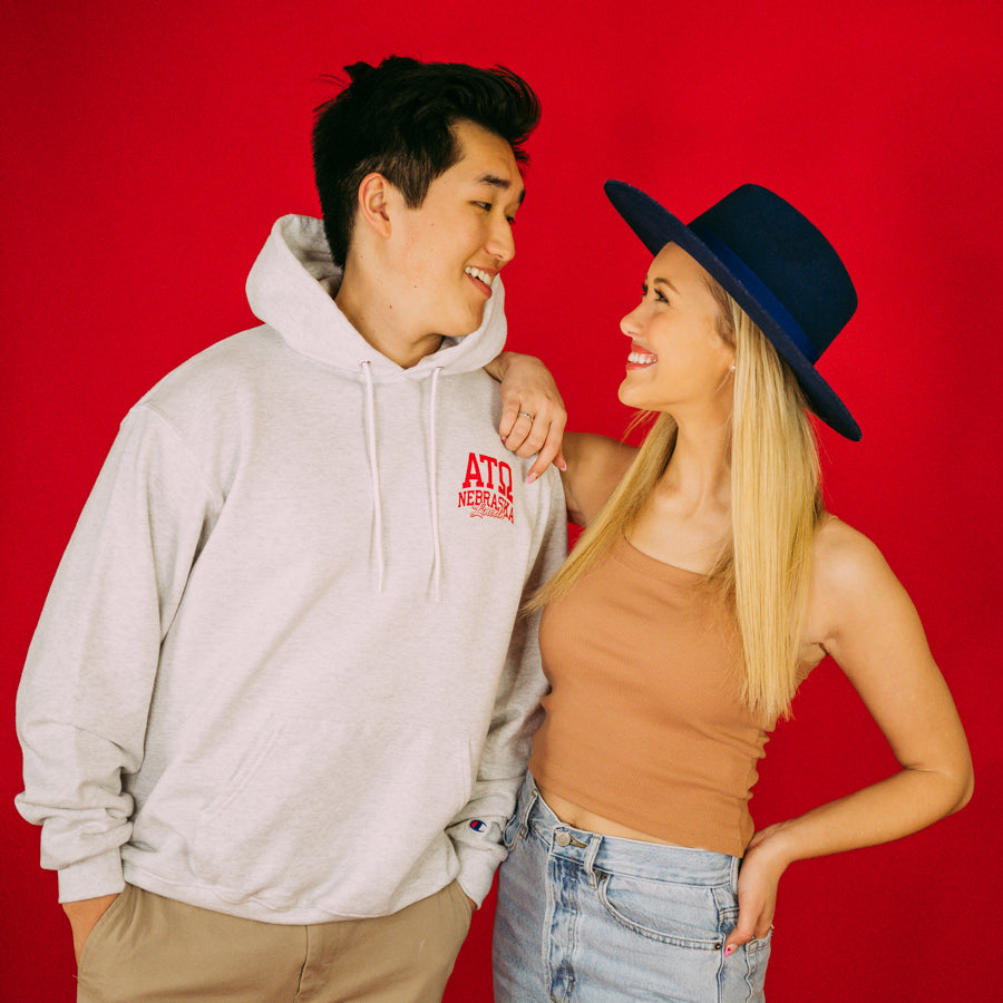Girl and Boy stand in front of a red backdrop. Boy wears a gray hoodie with Alpha Tau Omega letters on the left corner.