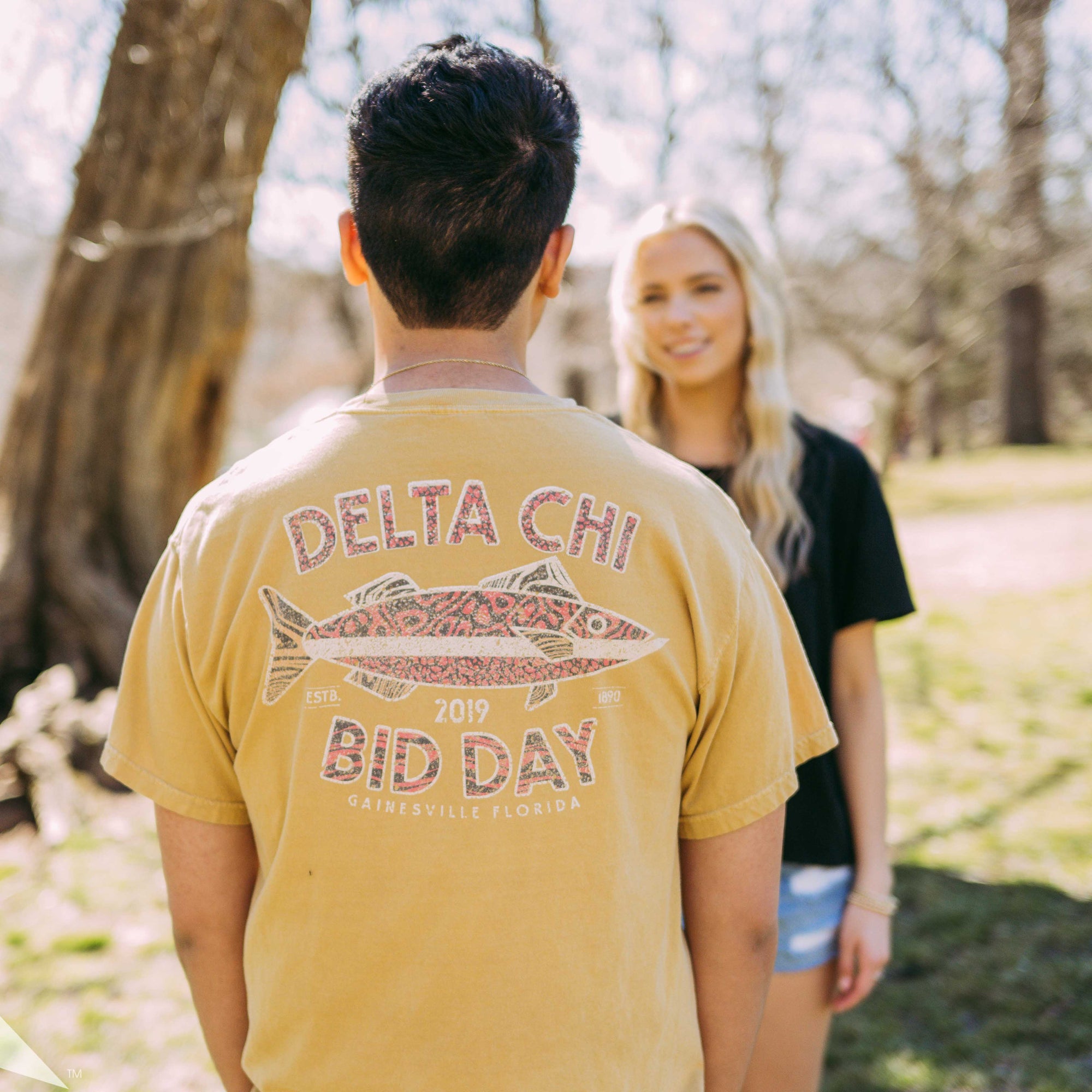 Boy with back facing the camera wearing a yellow Delta Chi tshirt with a picture of a fish on it.
